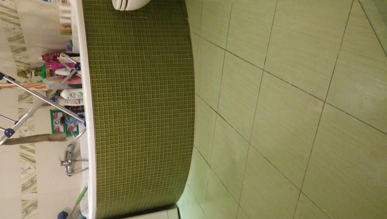 Zielona mozaika na obudowie wanny sąsiadująca z większymi, kwadratowymi, jasnymi płytkami podłogowymi w łazience, z widocznym kranem i suszarką do ubrań.