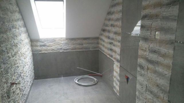 Łazienka w trakcie remontu, widoczne częściowo wykończone ściany z szarej, strukturalnej okładziny imitującej kamień, gładkie szare płytki na dolnej części ścian i podłodze, okno dachowe w skosie...