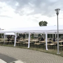 Białe namioty plenerowe z drewnianymi stołami i ławkami, ludzie siedzący przy stołach na zewnątrz.