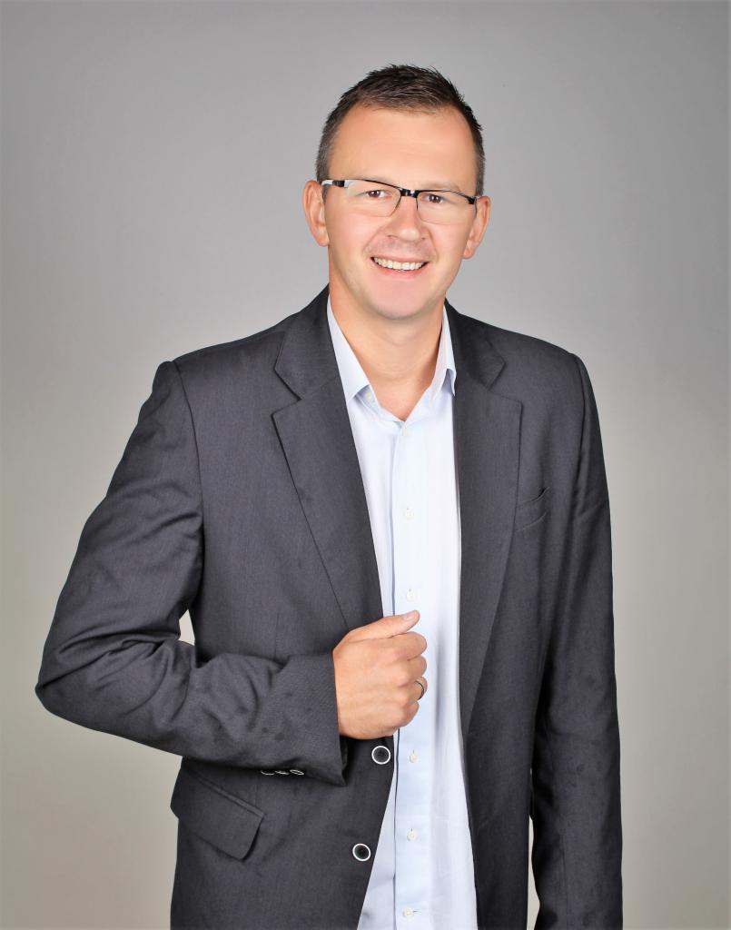 Portret uśmiechniętego mężczyzny w okularach i szarym garniturze na jednolitym tle. Widoczny od pasa w górę, z ręką na klapie marynarki.