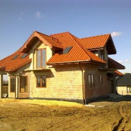 Dom jednorodzinny w trakcie budowy, z widoczną czerwoną dachówką, murem z cegły dziurawki i oknami z brązowymi ramami, otoczony ziemią.