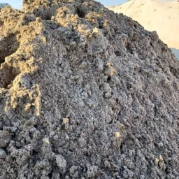 Duża pryzma szarego, wilgotnego piasku budowlanego w promieniach popołudniowego słońca, z widocznym w tle jaśniejszym nasypem i błękitnym niebem.