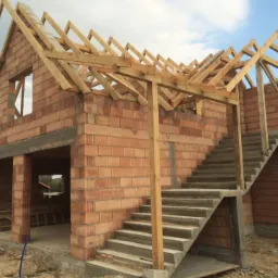 Surowy, dwukondygnacyjny budynek z czerwonej cegły w trakcie budowy, widoczne drewniane elementy konstrukcji dachu i betonowe schody zewnętrzne, otoczenie placu budowy.