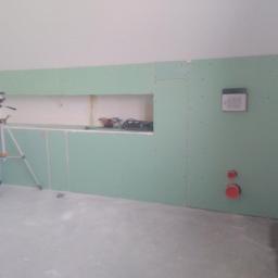 Maxpol - Wnętrze w trakcie montażu płyt kartonowo-gipsowych, widoczny poziomica laserowa na statywie, otwór w ścianie, instalacja wodna i elektryczna.