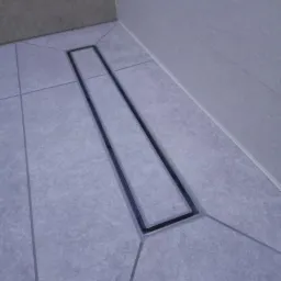 Szary odpływ liniowy wkomponowany w jasnoszare płytki podłogowe i ścienne w narożniku łazienki, z delikatnym spadkiem płytek w kierunku odpływu.