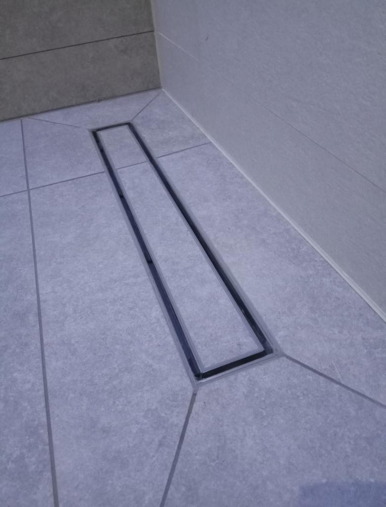 Szary odpływ liniowy wkomponowany w jasnoszare płytki podłogowe i ścienne w narożniku łazienki, z delikatnym spadkiem płytek w kierunku odpływu.