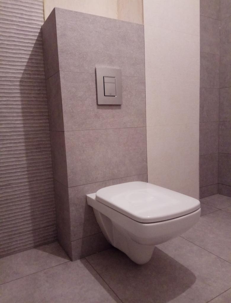 Wisząca toaleta z podtynkową spłuczką na tle szarych płytek z delikatną fakturą i białych płytek, widoczne szare płytki podłogowe.