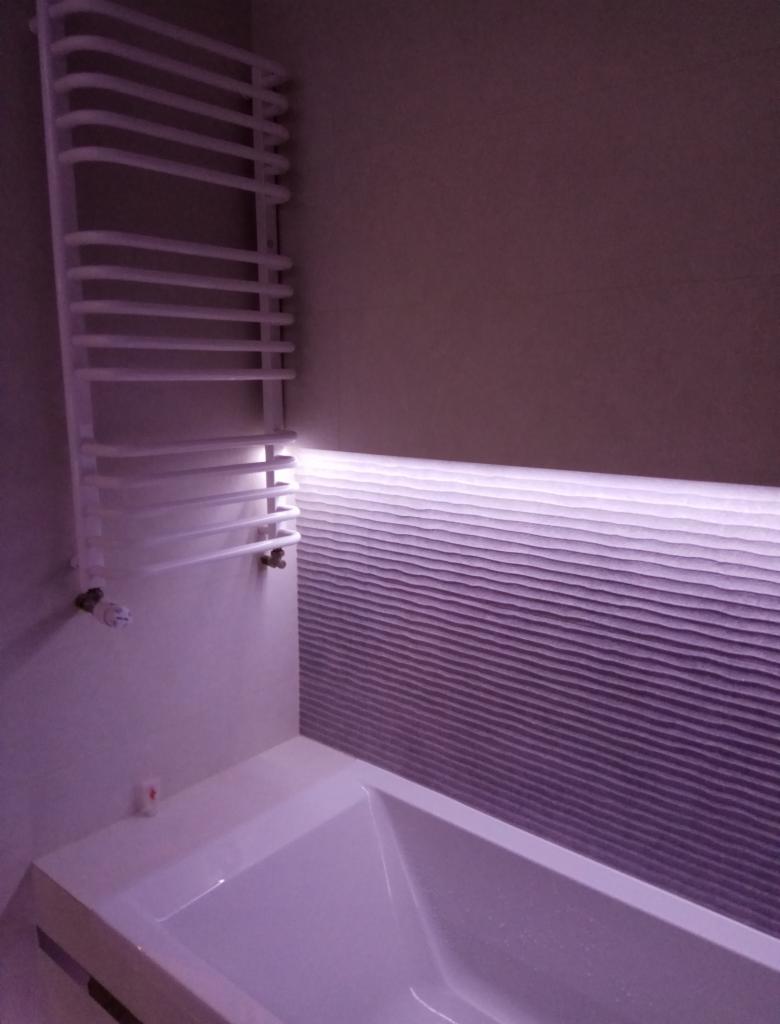 Nowoczesna łazienka z białą wanną, grzejnikiem drabinkowym i podświetlaną, teksturowaną ścianą.