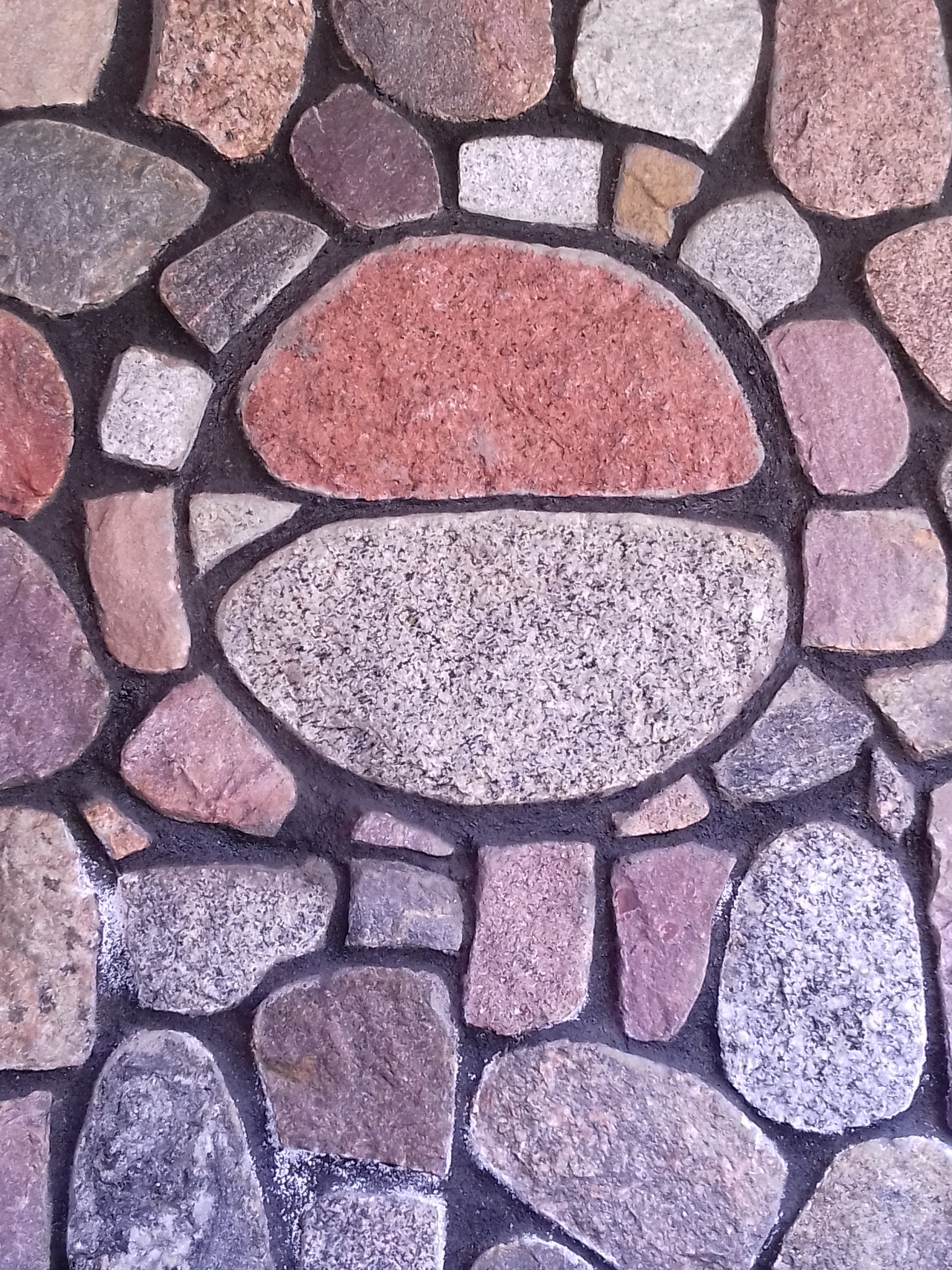 Dekoracyjna ściana z nieregularnych kamieni w odcieniach szarości, różu i bieli, ułożonych w mozaikę z ciemną fugą, tworzących unikalną teksturę.