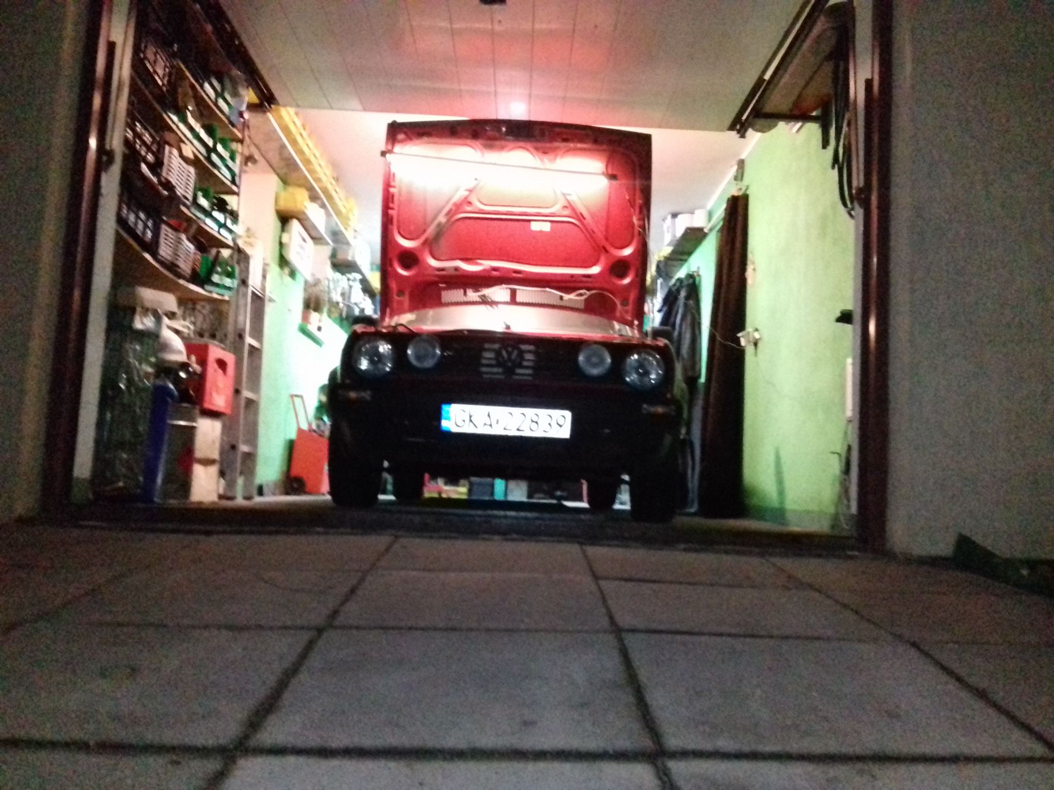 Czerwony samochód z otwartą maską w garażu, widok z niskiej perspektywy, oświetlenie sufitowe. Warsztat samochodowy z półkami w tle.