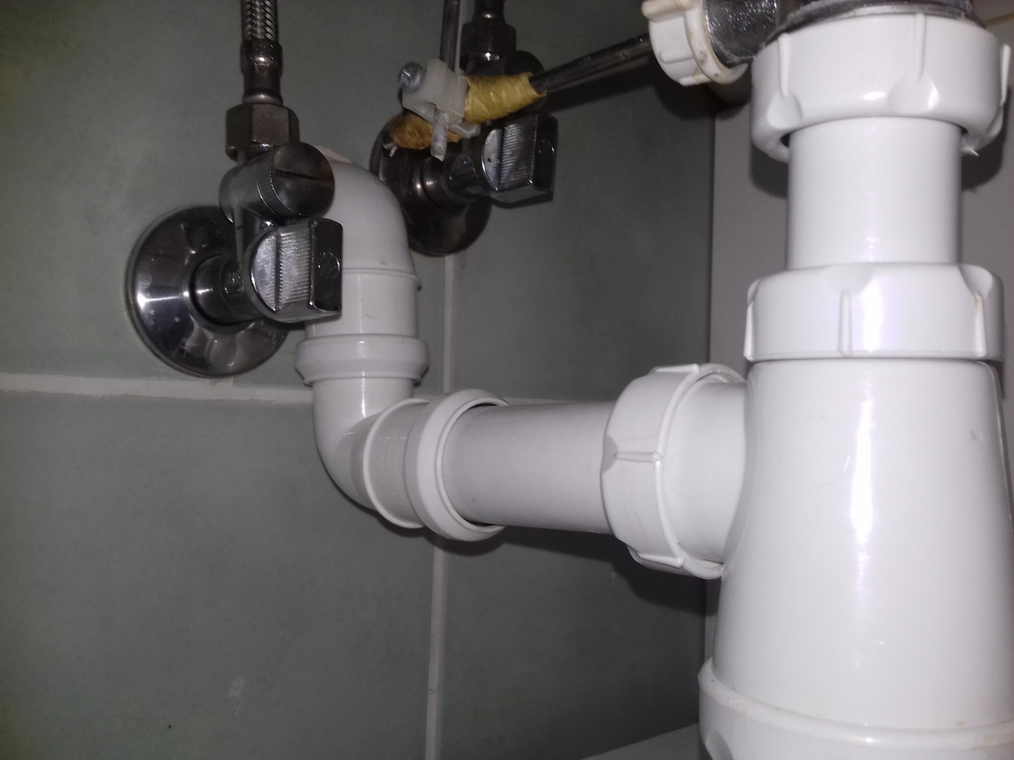 Biały syfon umywalkowy z metalowymi zaworami przy szarej ścianie. Widoczne łączenia rur i elementy instalacji wodnej. Ujęcie z bliska, brak szumów.