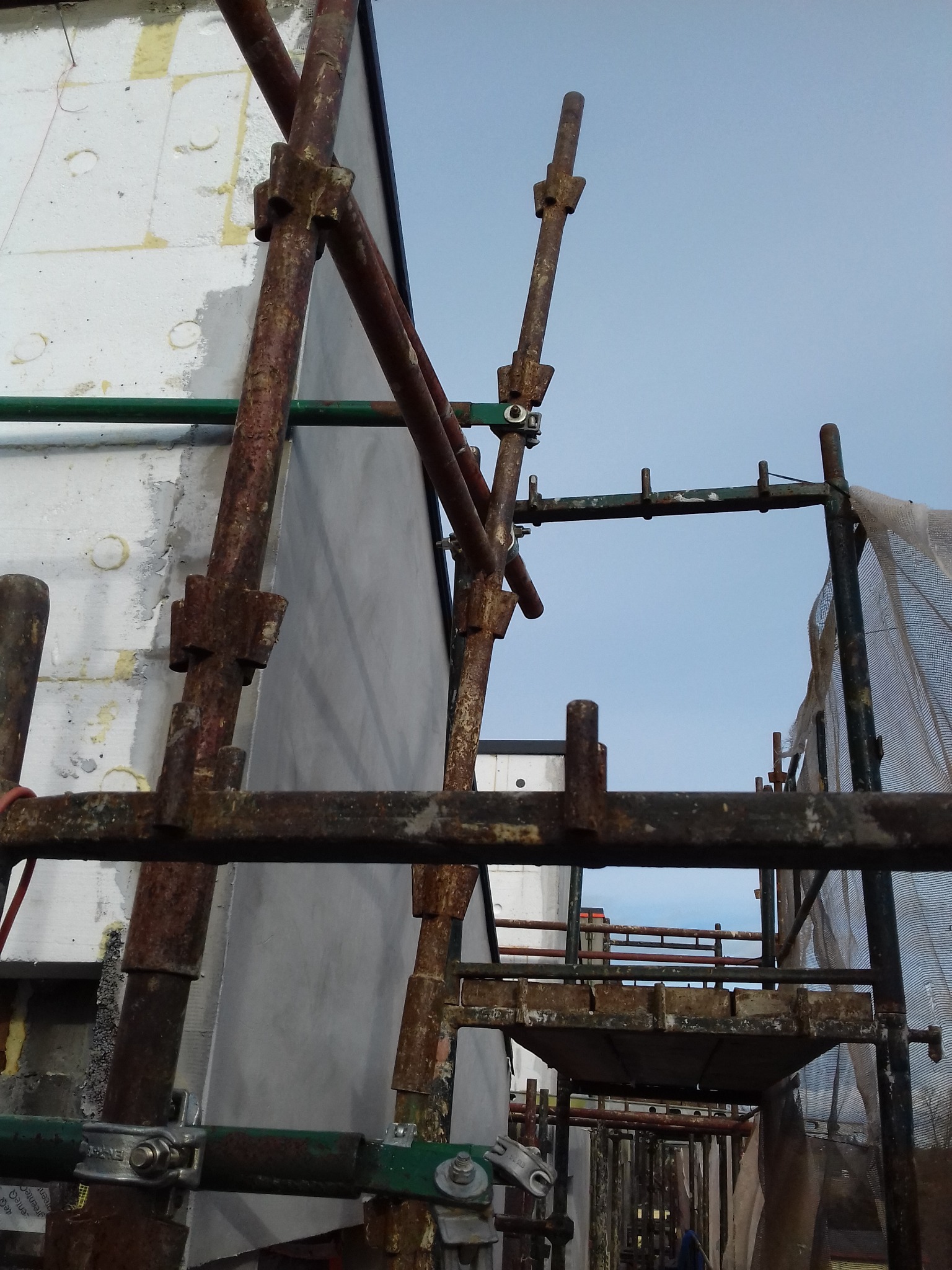 Ocieplanie budynku z rusztowaniem: widok z dołu na konstrukcję z rur, ocieploną ścianę i błękitne niebo. Elementy rusztowania zardzewiałe, co dodaje charakteru.