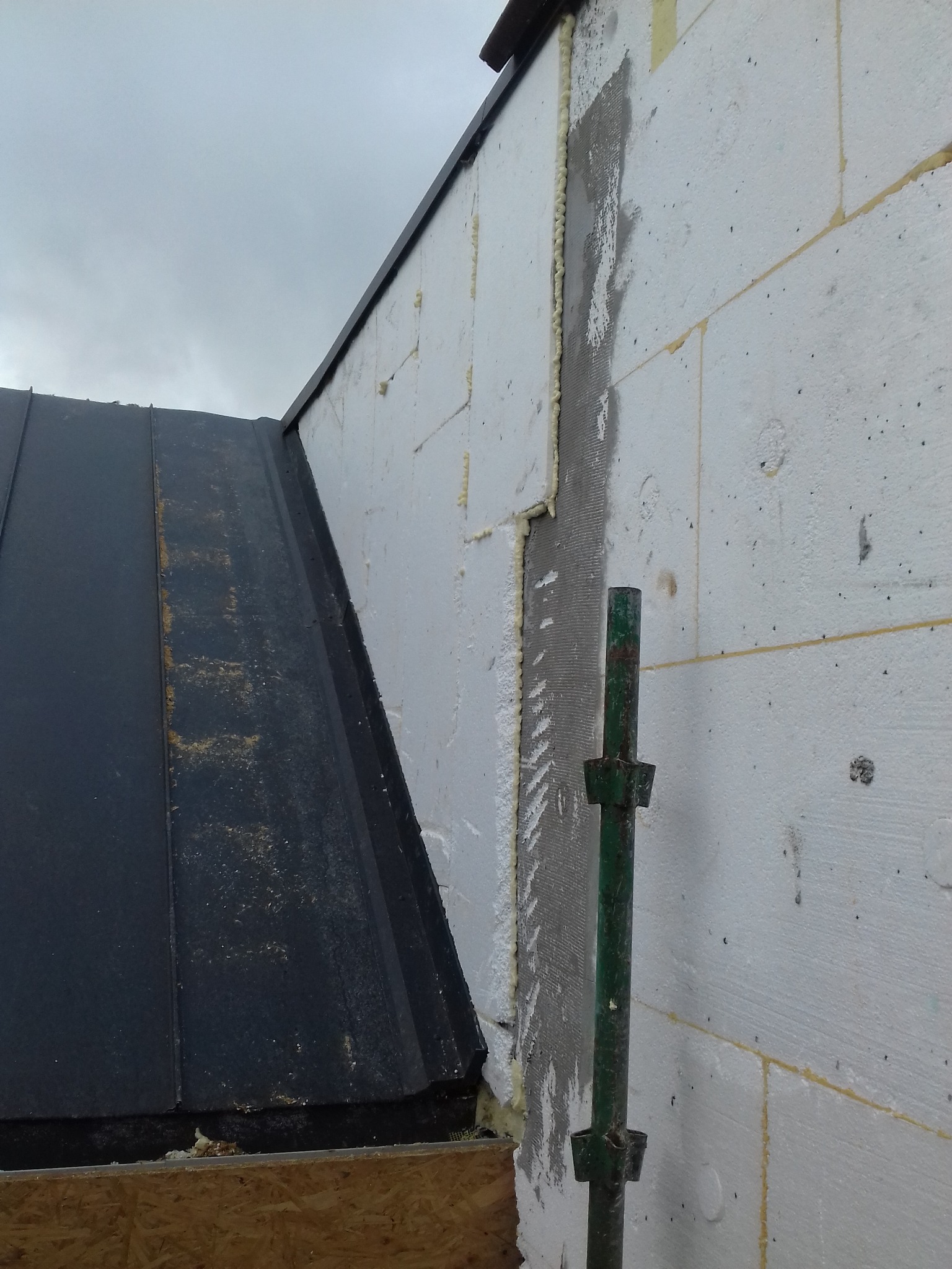 Ocieplenie ściany zewnętrznej z użyciem styropianu i siatki z klejem. Widoczne rusztowanie i fragment dachu pokrytego blachą trapezową.