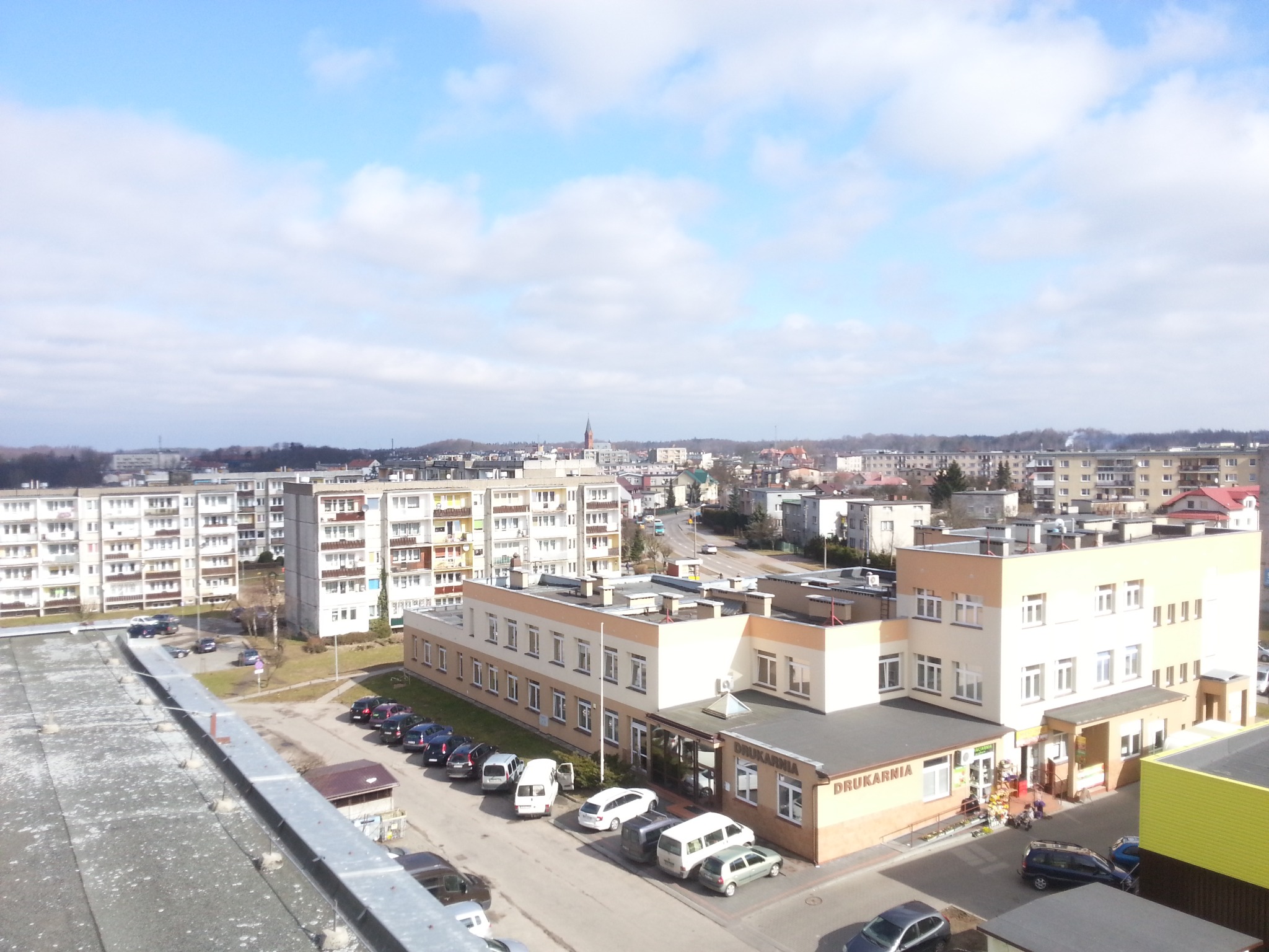 Panorama miasta z widokiem na budynek drukarni z napisem 'DRUKARNIA' na dachu, zaparkowane samochody i bloki mieszkalne w tle, pod błękitnym niebem z chmurami.