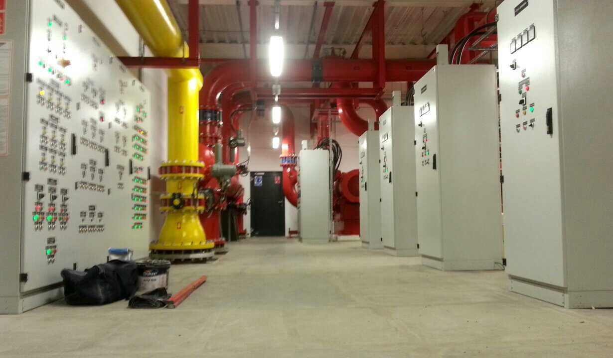 Instalacja systemów przeciwpożarowych: żółta rura, czerwone rury i białe szafy sterownicze w pomieszczeniu technicznym z oświetleniem. Widoczny sprzęt roboczy.