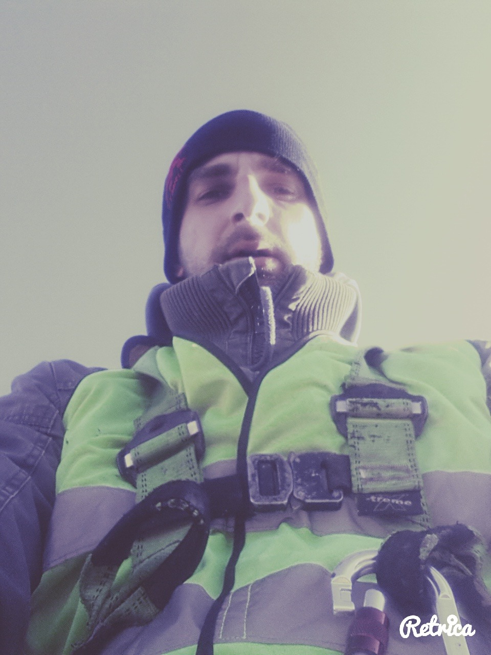 Autoportret mężczyzny w czapce i odzieży roboczej z uprzężą, z perspektywy żabiej, na jasnym tle. Widoczny fragment twarzy i górna część ciała.