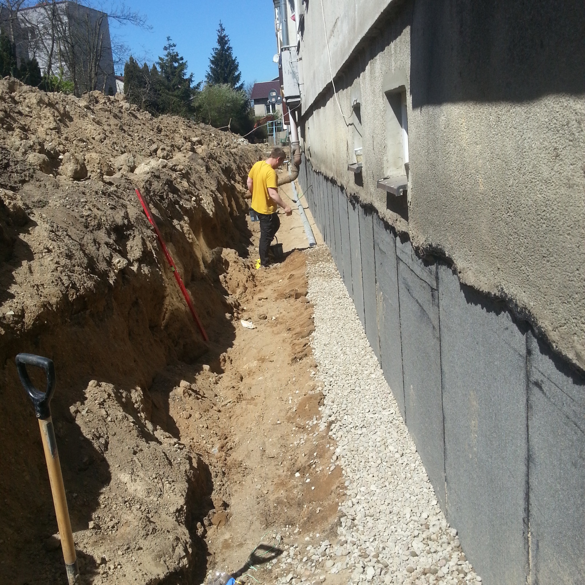 Odkryte fundamenty budynku z hydroizolacją, drenażem i wykopem. Widoczny pracownik w żółtej koszulce. Prace ziemne i izolacyjne w toku.