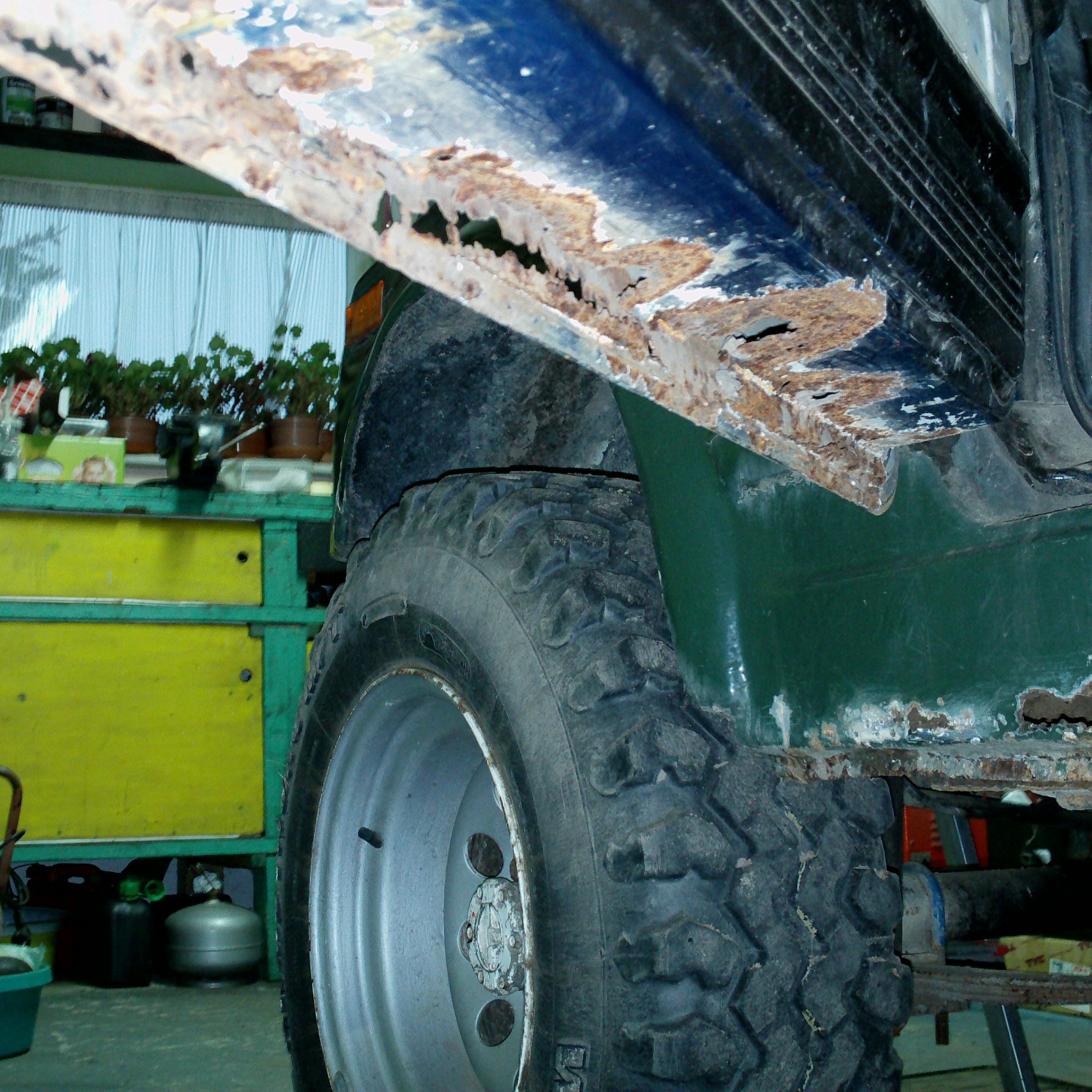 Zardzewiały fragment karoserii samochodu terenowego przy oponie. Widoczne ogniska korozji i perforacje blachy. W tle warsztat z narzędziami i roślinami.