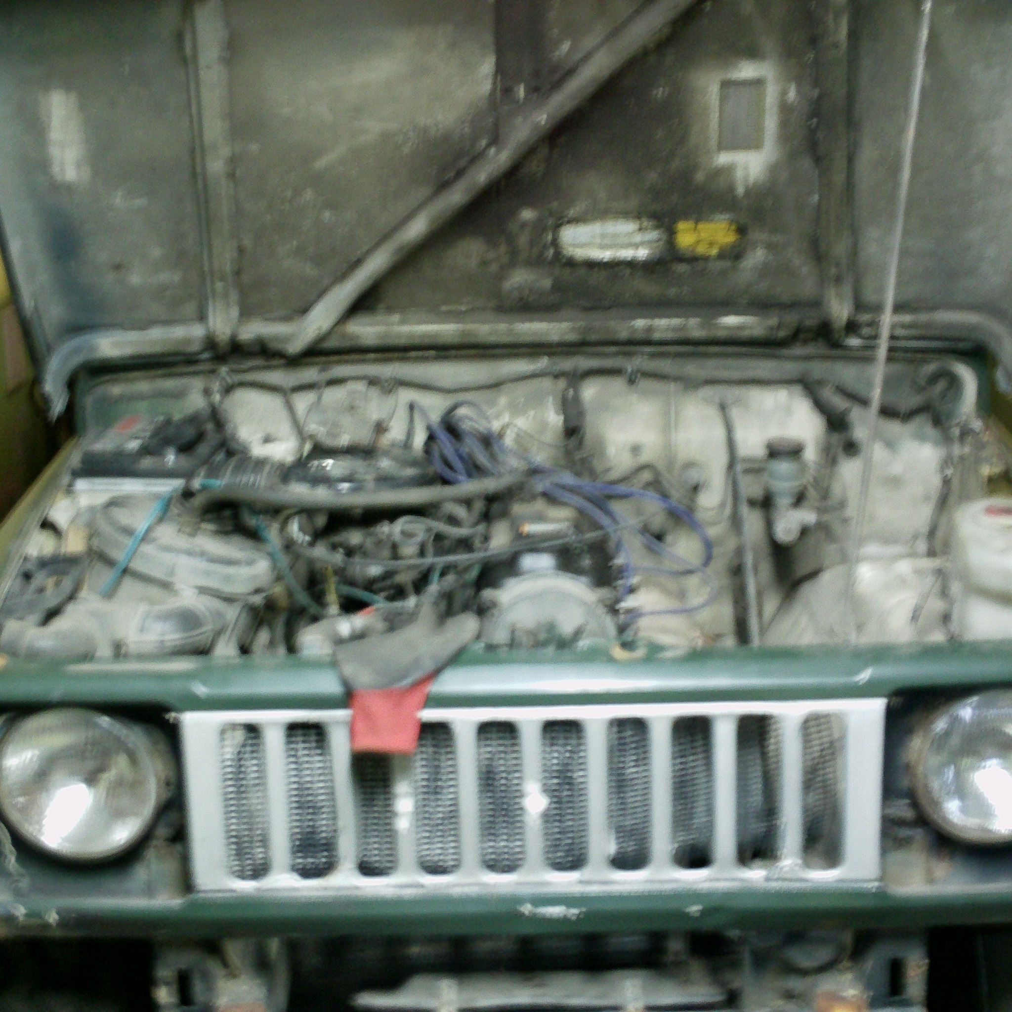 Otwarta maska zielonego samochodu terenowego, widoczny silnik z przewodami, akumulator i płyny eksploatacyjne. Na masce widoczne naklejki.