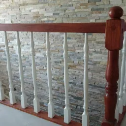 Fragment schodów z drewnianą balustradą z białymi tralkami na tle ściany wyłożonej kamieniem dekoracyjnym. Widoczne narzędzia i obuwie na podłodze sugerują trwający montaż.