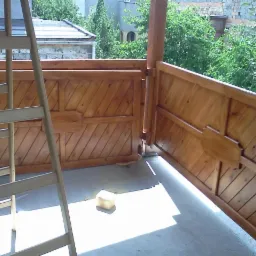 W trakcie montażu drewnianej balustrady na tarasie, widoczna drabina i fragmenty desek z ukośnym wzorem, jasne drewno, betonowa podłoga, zieleń w tle.