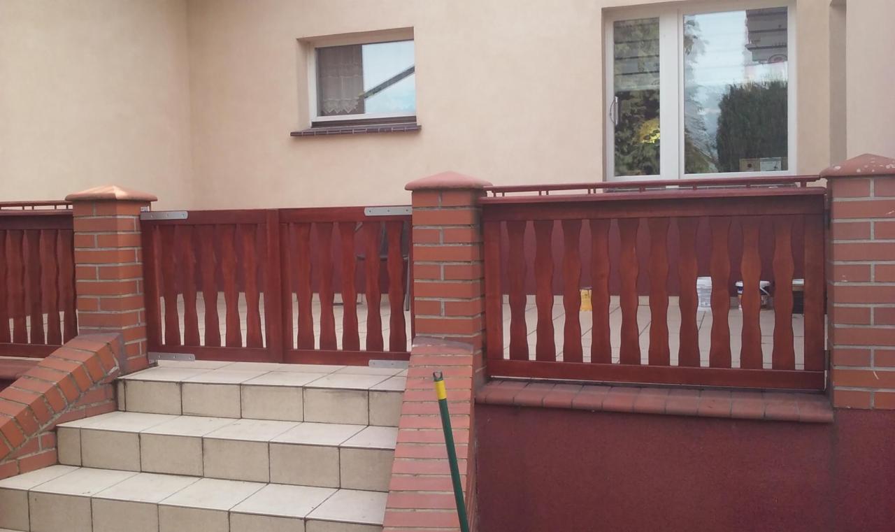 Wejście do domu z niskimi schodami wyłożonymi płytkami i balustradą z ciemnego drewna z pionowymi szczeblami, osadzoną na ceglanych słupkach.