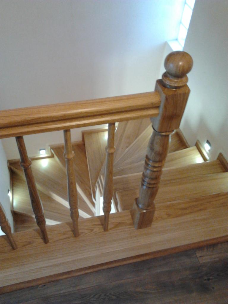 Widok z góry na kręte schody z jasnego drewna z drewnianą balustradą z okrągłym zakończeniem słupka, oświetlone małymi lampkami LED w ścianie.