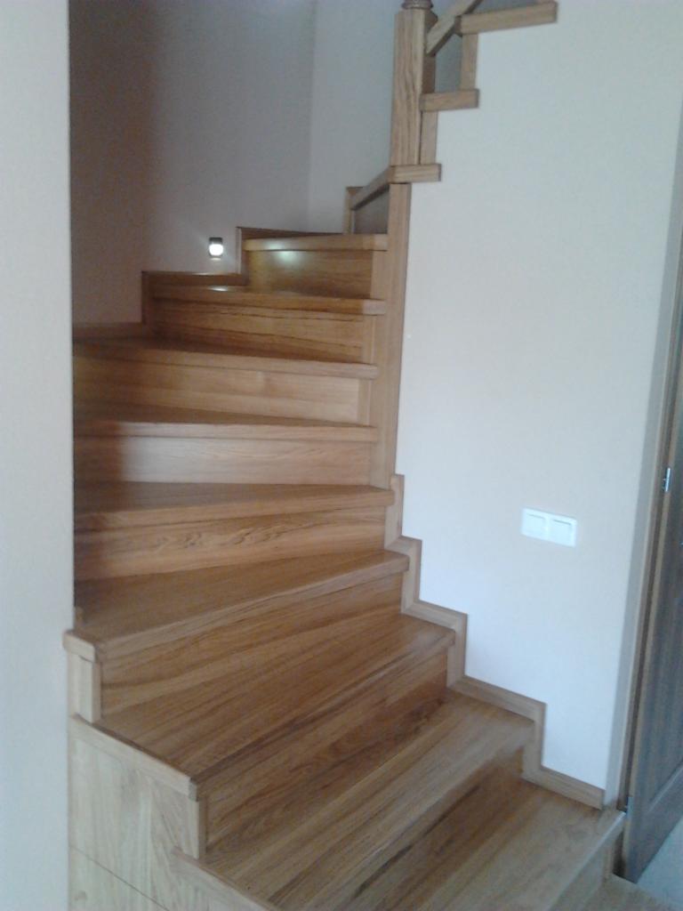 Jasne, drewniane schody z podświetleniem stopnia i prostą balustradą przy ścianie w jasnym wnętrzu.