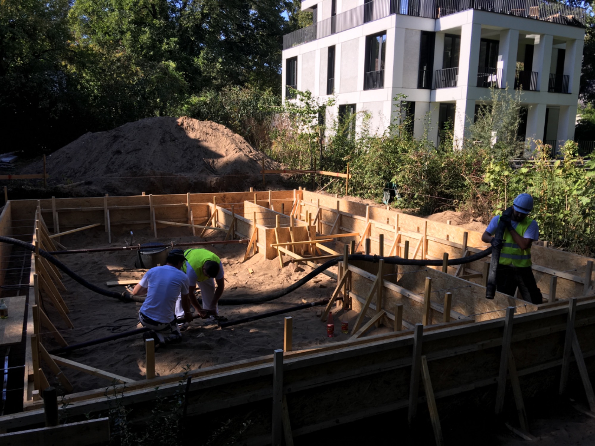 Przygotowanie szalunku pod fundamenty domu na tle sterty ziemi i nowoczesnego budynku, dwóch pracowników obsługuje pompę do betonu, trzeci w kasku nadzoruje prace.