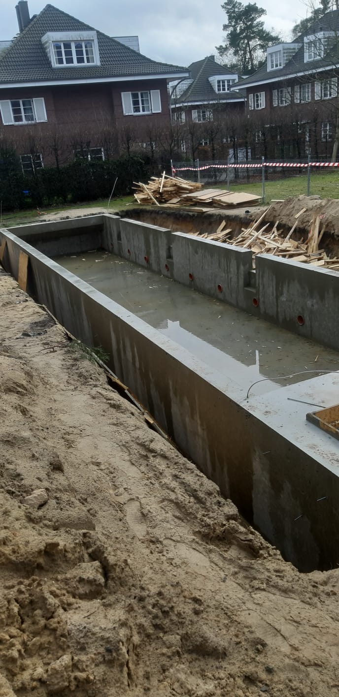 Wylany betonowy fundament pod budowę basenu, widoczne otwory na instalacje, w tle domy jednorodzinne i stosy drewna budowlanego.