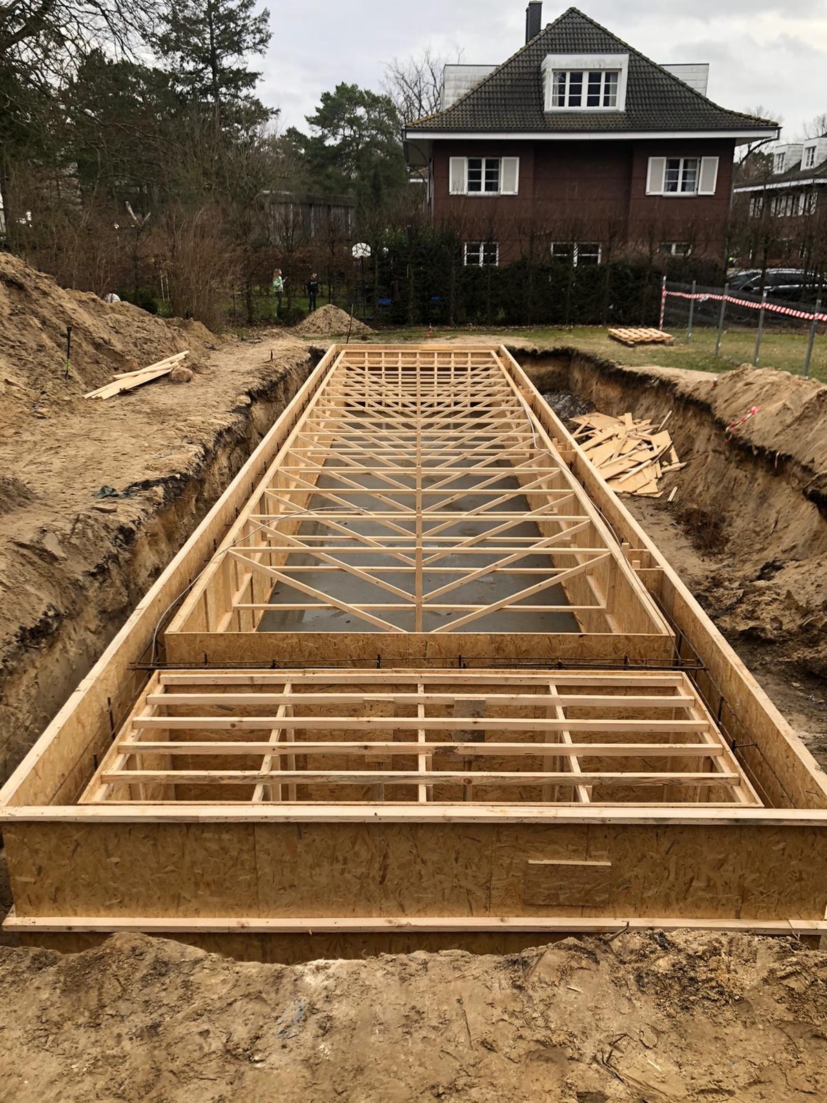 Szeroki wykop pod budowę z drewnianym szalunkiem i zbrojeniem, widok z góry na przygotowany teren pod wylewkę fundamentów domu jednorodzinnego na tle budynku mieszkalnego.