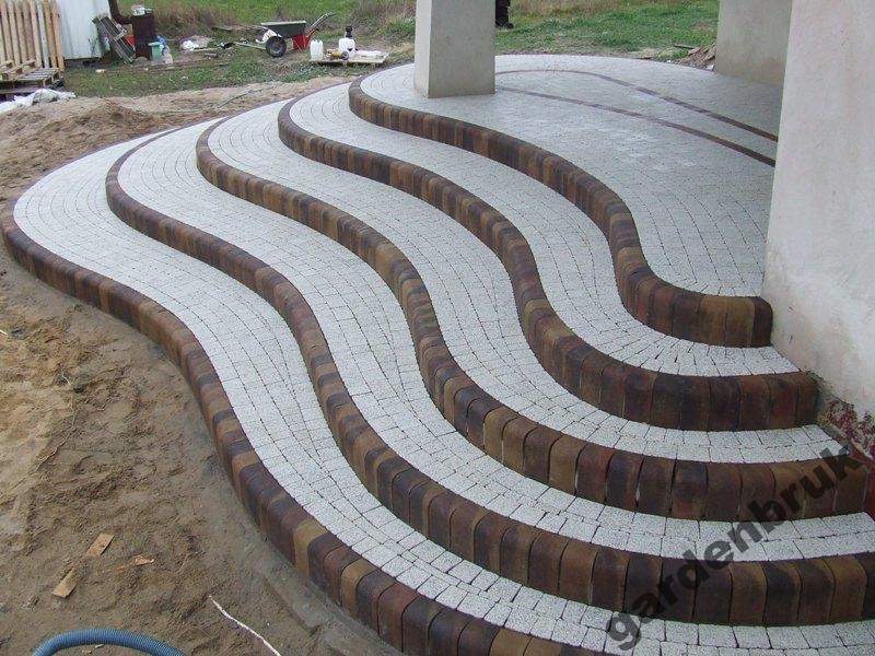 Faliste schody wejściowe ułożone z jasnej kostki brukowej obramowanej ciemniejszą kostką, prowadzące do budynku z kolumną.