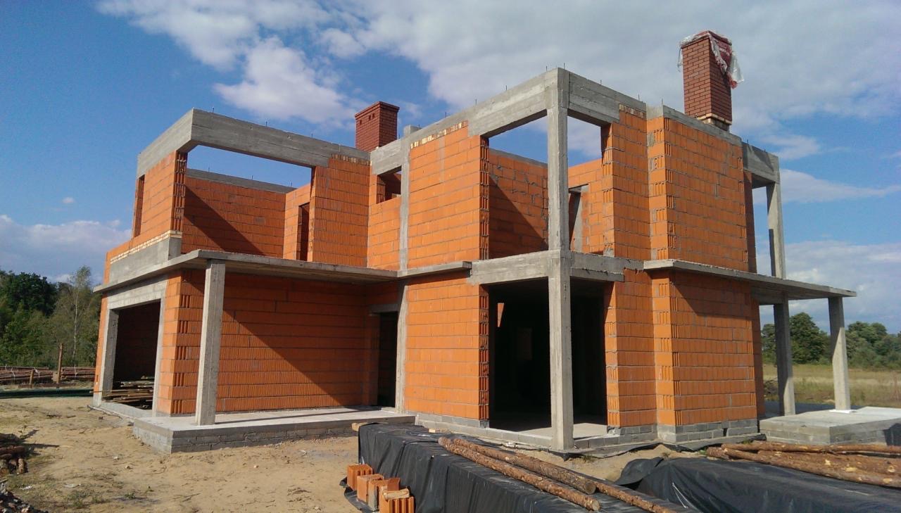 Dwukondygnacyjny dom w stanie surowym otwartym z czerwonej cegły, betonowe nadproża i słupy, widoczne kominy z cegły, teren budowy z drewnem i czarną folią, błękitne niebo w tle.