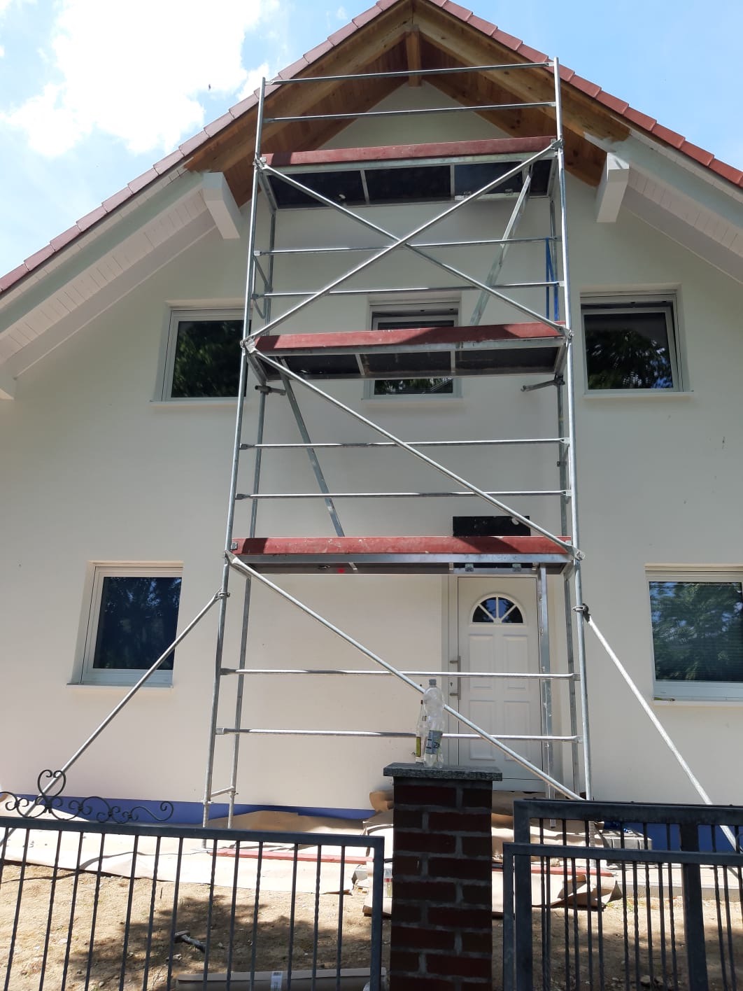 Biały dom jednorodzinny w trakcie tynkowania elewacji, widoczne rusztowanie, fragment ogrodzenia i butelka wody na ceglanym słupku.