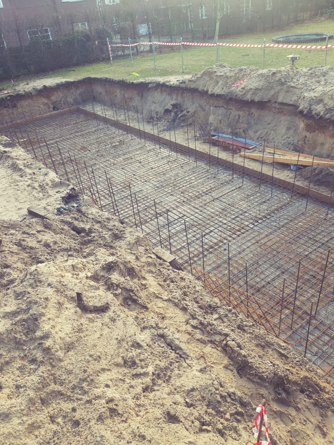 Przygotowany wykop z ułożonym zbrojeniem stalowym na piasku, zabezpieczony taśmą ostrzegawczą w tle.