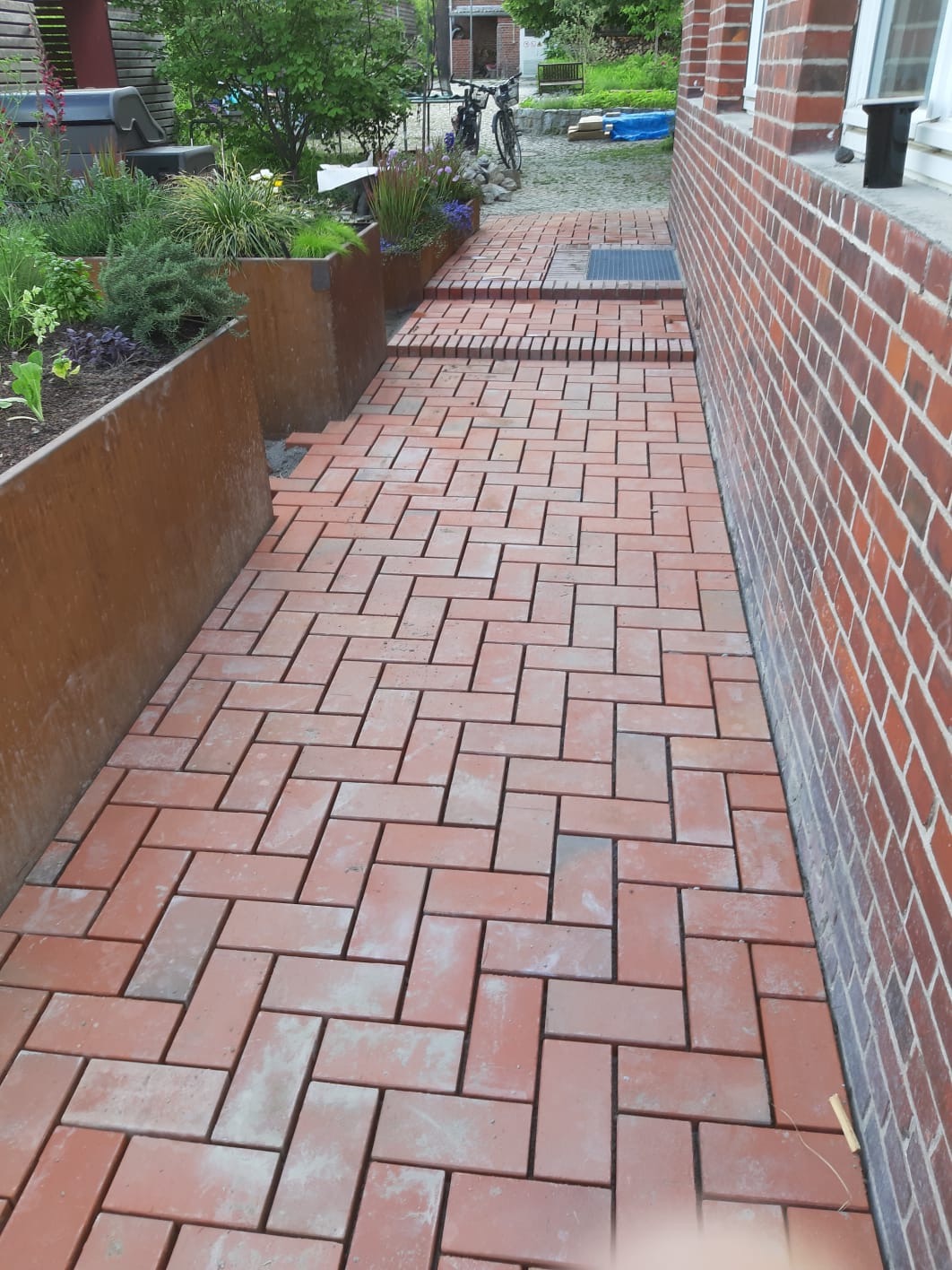 Czerwona ścieżka z kostki ułożona w jodełkę prowadząca przez ogród, z metalowymi donicami z roślinami po jednej stronie i ceglaną ścianą budynku po drugiej. Schodki z kostki brukowej prowadzą w górę.