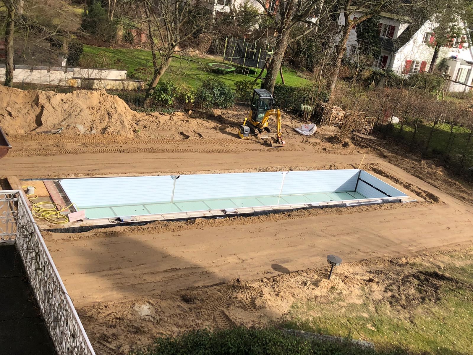 Wykopany dół pod basen z widoczną konstrukcją ścian basenu, teren wyrównany, w tle mini koparka i domy.