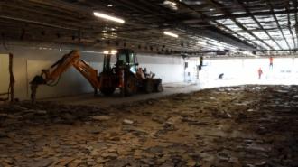 Wnętrze hali sklepu Lidl w trakcie rozbiórki. Pomarańczowa koparko-ładowarka przy ścianie, gruz na podłodze, widoczne elementy konstrukcji stropu i oświetlenie. Pracownicy w oddali.