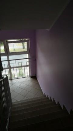 Klatka schodowa w bloku mieszkalnym po odświeżeniu: pomalowane ściany na fioletowo, jasne schody z ciemnymi fugami, okno z metalową barierką z widokiem na zieleń.