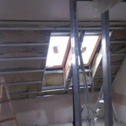 Montaż izolacji termicznej i metalowej konstrukcji pod zabudowę poddasza z oknami dachowymi, widoczna drewniana drabina.