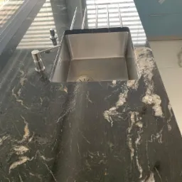 Czarny, wzorzysty blat kuchenny z granitu z wbudowanym, prostokątnym zlewem ze stali nierdzewnej i nowoczesną baterią, oświetlony przez żaluzje okienne.