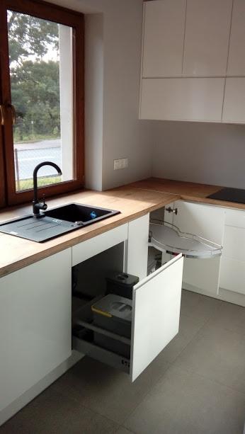 Nowoczesna kuchnia z białymi szafkami, drewnianym blatem i otwartą szufladą z pojemnikami na segregację śmieci; widoczny zlew z czarną baterią i narożna szafka z półkami obrotowymi.