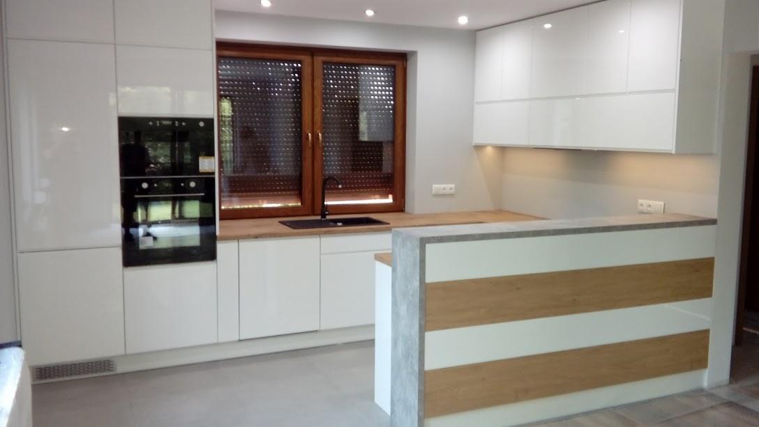 Nowoczesna, biała kuchnia z drewnianym blatem i oknem z żaluzjami, widok na zabudowaną piekarnik i wyspę z dekoracyjnymi panelami.