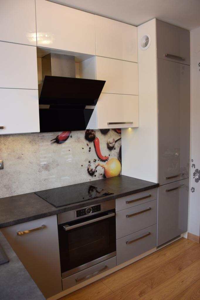 Nowoczesna kuchnia z białymi i szarymi szafkami, czarnym okapem i panelem szklanym z motywem warzyw, oświetlona punktowo pod szafkami.