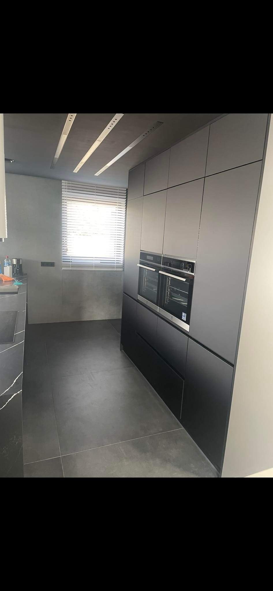Minimalistyczna, szara kuchnia z zabudową sięgającą sufitu, z dwoma piekarnikami wkomponowanymi w szafki, betonowa ściana i podłoga z szarych płytek, oświetlenie sufitowe listwowe.