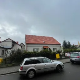 Dom jednorodzinny z nowym, czerwonym dachem we Wschowie, widoczny z ulicy, z zaparkowanymi samochodami i żółtą skrzynką gazową.