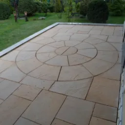 Ułożony taras z piaskowej kostki z dekoracyjnym wzorem koła w centrum, obrzeże z szarego granitu, trawnik w tle.