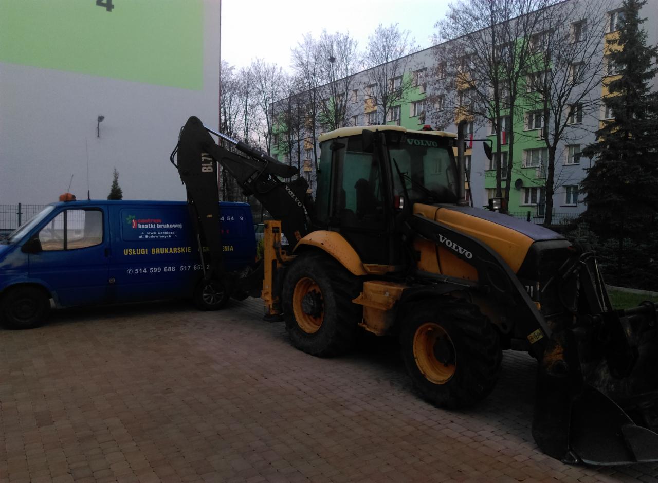 Żółto-czarna koparko-ładowarka Volvo BL71 stoi na kostce brukowej obok niebieskiego busa z reklamą usług brukarskich na tle bloku mieszkalnego.