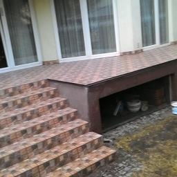 Remont mieszkan - Wyłożony płytkami taras z niskimi schodami i ukrytym pod tarasem miejscem do przechowywania, widoczne wiadra i zwinięty wąż ogrodowy.