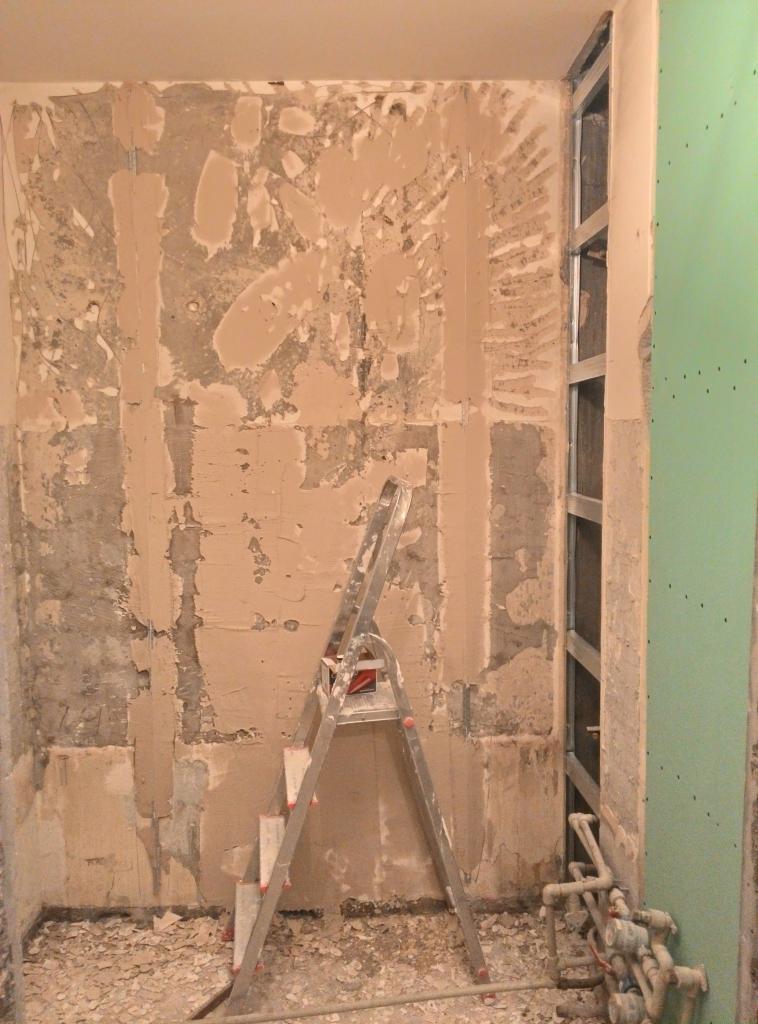 Etap przygotowania ściany w łazience do dalszych prac wykończeniowych, widoczne ślady po skuciu starych płytek, częściowo nałożona nowa warstwa zaprawy, metalowa drabina oparta o ścianę, fragment...